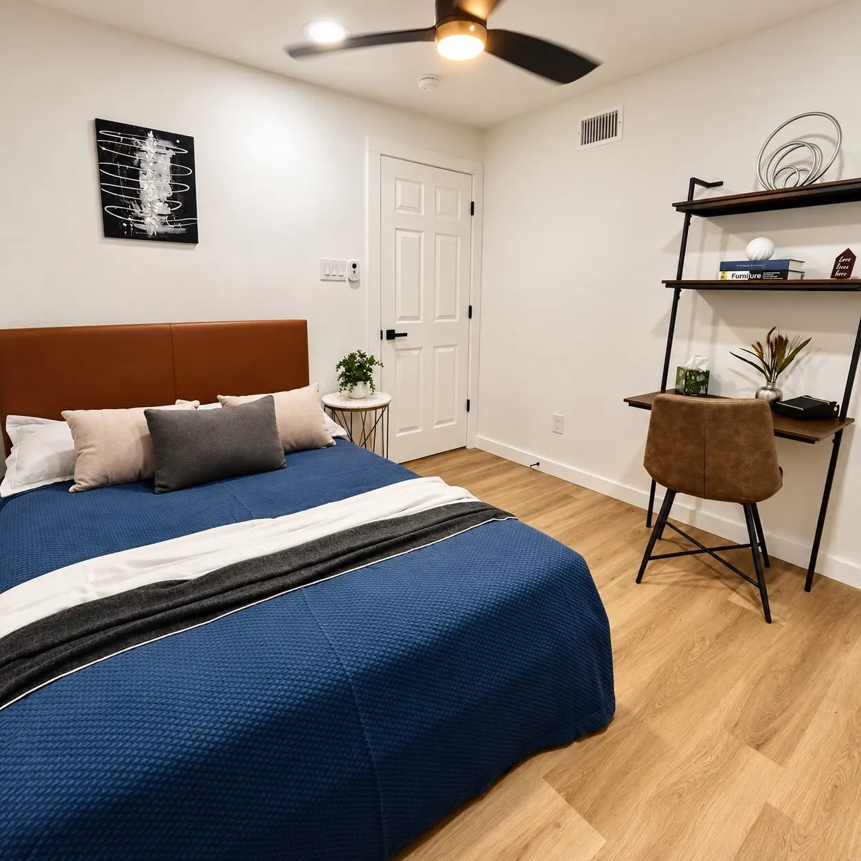 Wood-finish flooring, ceiling fan with integrated lighting, white paneled door with matte black hardware, and an integrated wall shelf with desk space