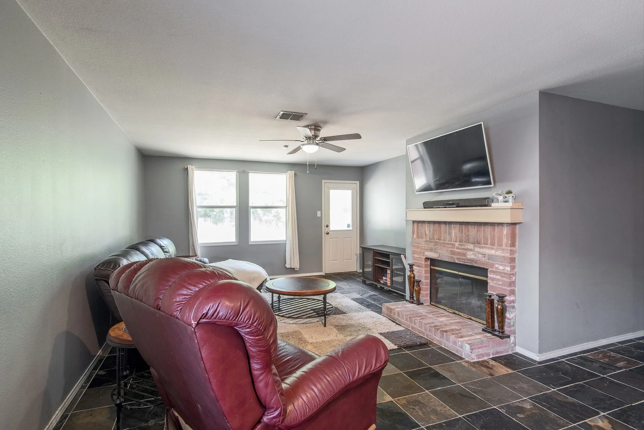 Living room featuring dark stone finish flooring, a brick fireplace, and a ceiling fan