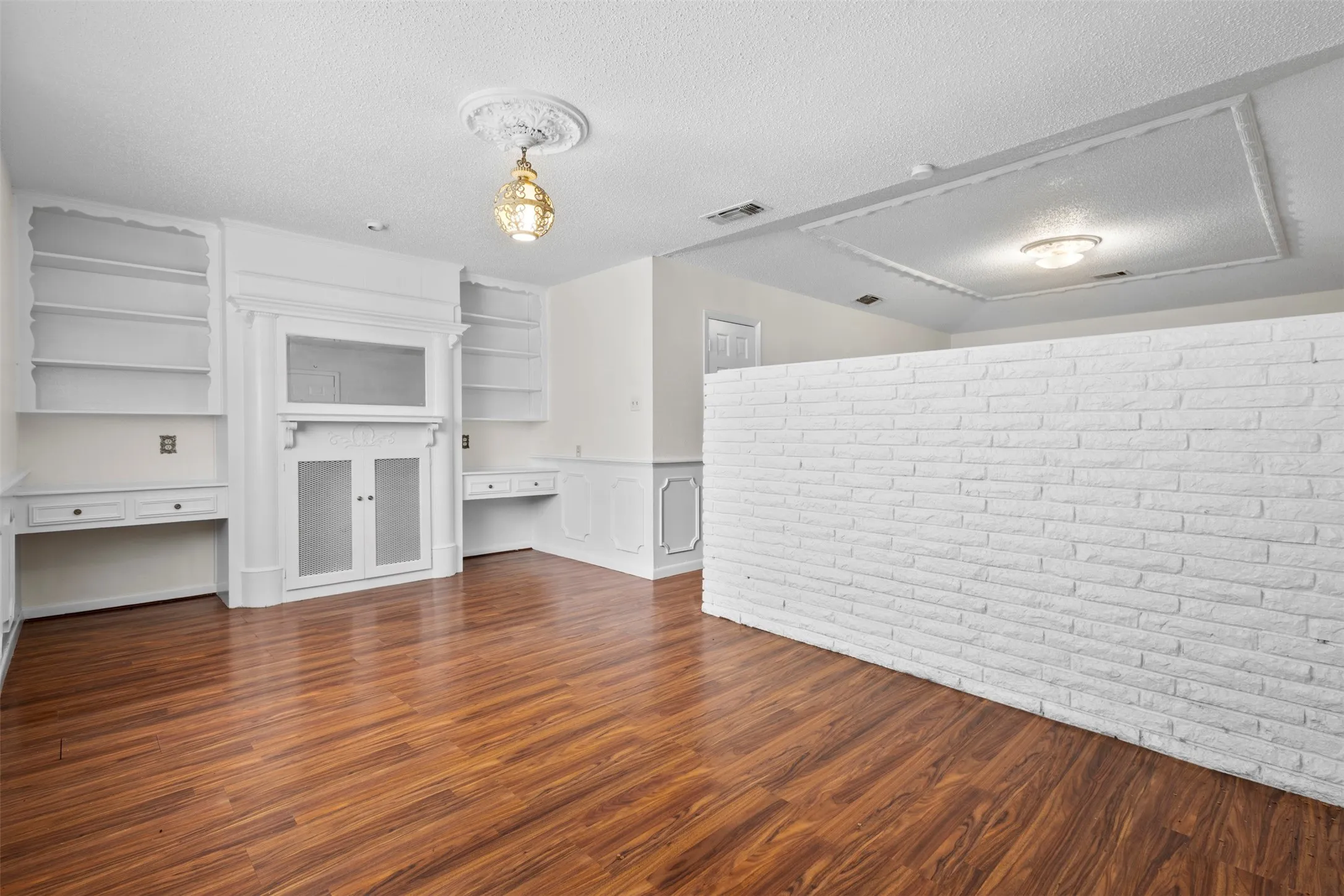 Unfurnished living room with built in desk, a textured ceiling, dark wood-style floors, brick wall, and a wainscoted wall