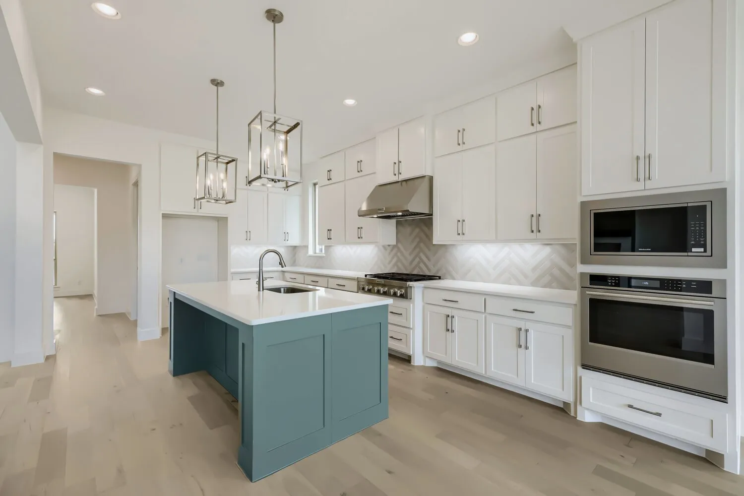 Modern kitchen featuring light wood-finish flooring, white upper and lower cabinetry, a blue-painted kitchen island with a white countertop, stainless steel appliances, and two hanging pendant light fixtures
