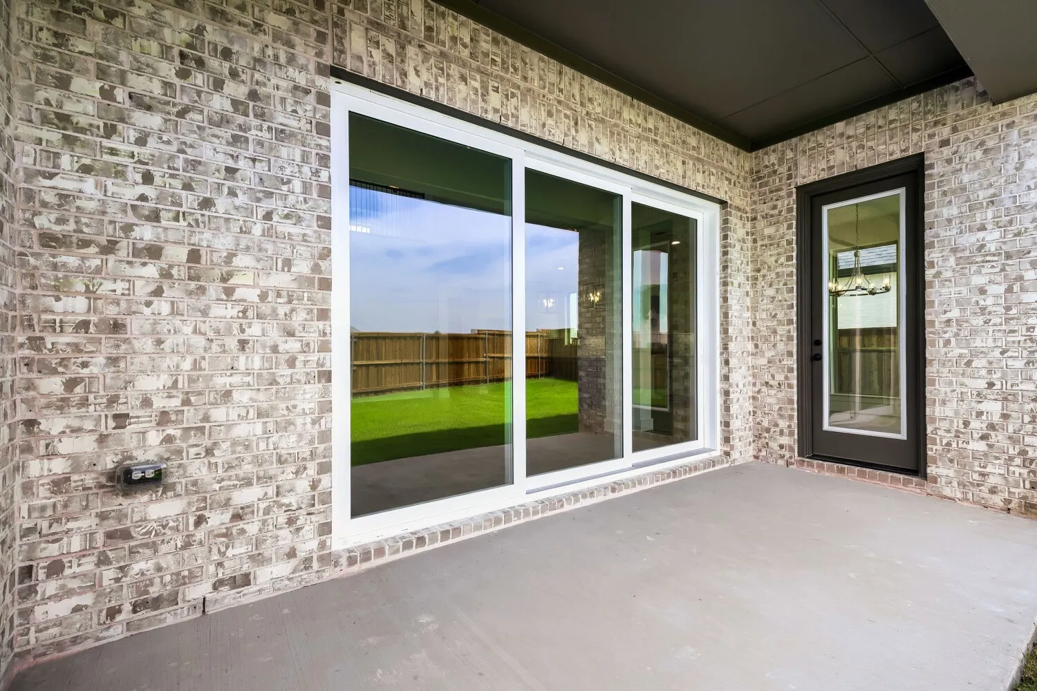 Outdoor patio featuring a gray concrete surface, light-toned brick exterior, white-framed sliding glass doors, a dark-framed glass-paneled entry door, and a dark soffit