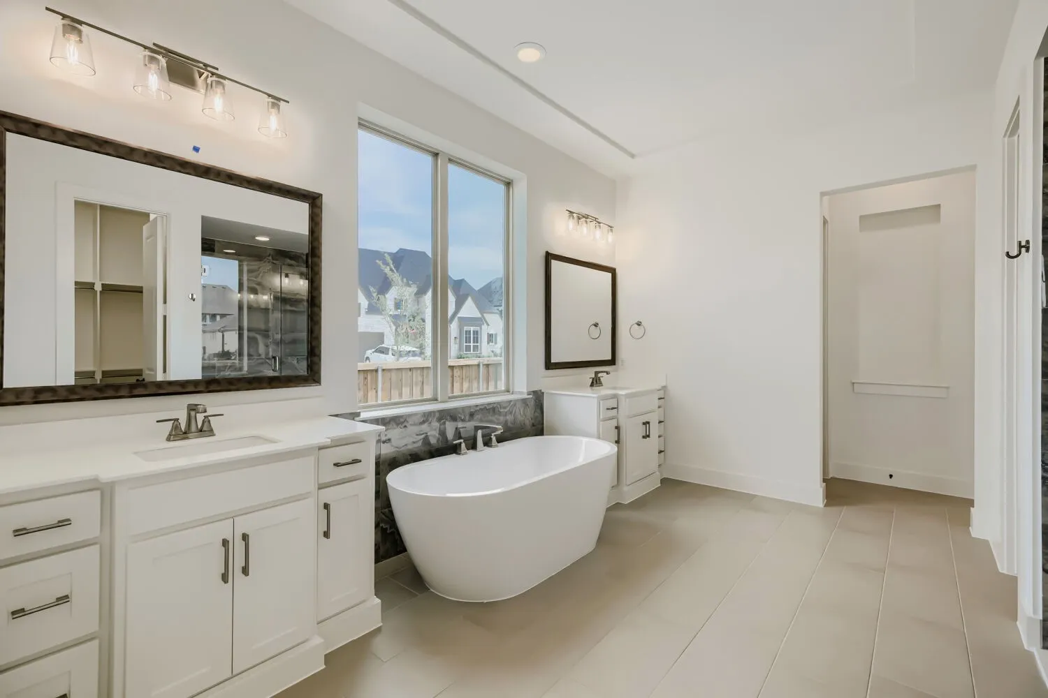 Spacious bathroom featuring a freestanding soaking tub, dual vanity with white cabinetry and integrated sinks, large window, and light-toned tile flooring