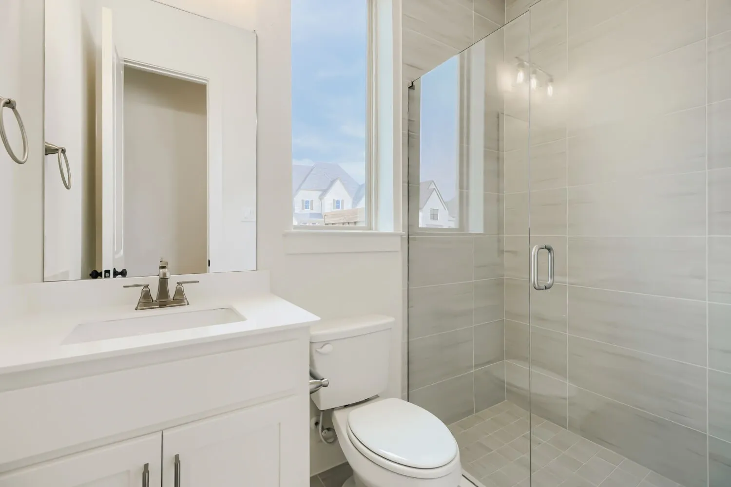 Contemporary bathroom featuring a frameless glass shower enclosure, large format tile shower surround, window with white trim, white vanity with integrated sink, and brushed nickel fixtures