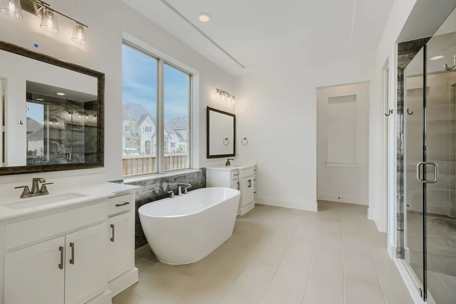 Freestanding soaking tub with dark stone surround, dual vanities with white cabinetry, large window, and glass-enclosed shower