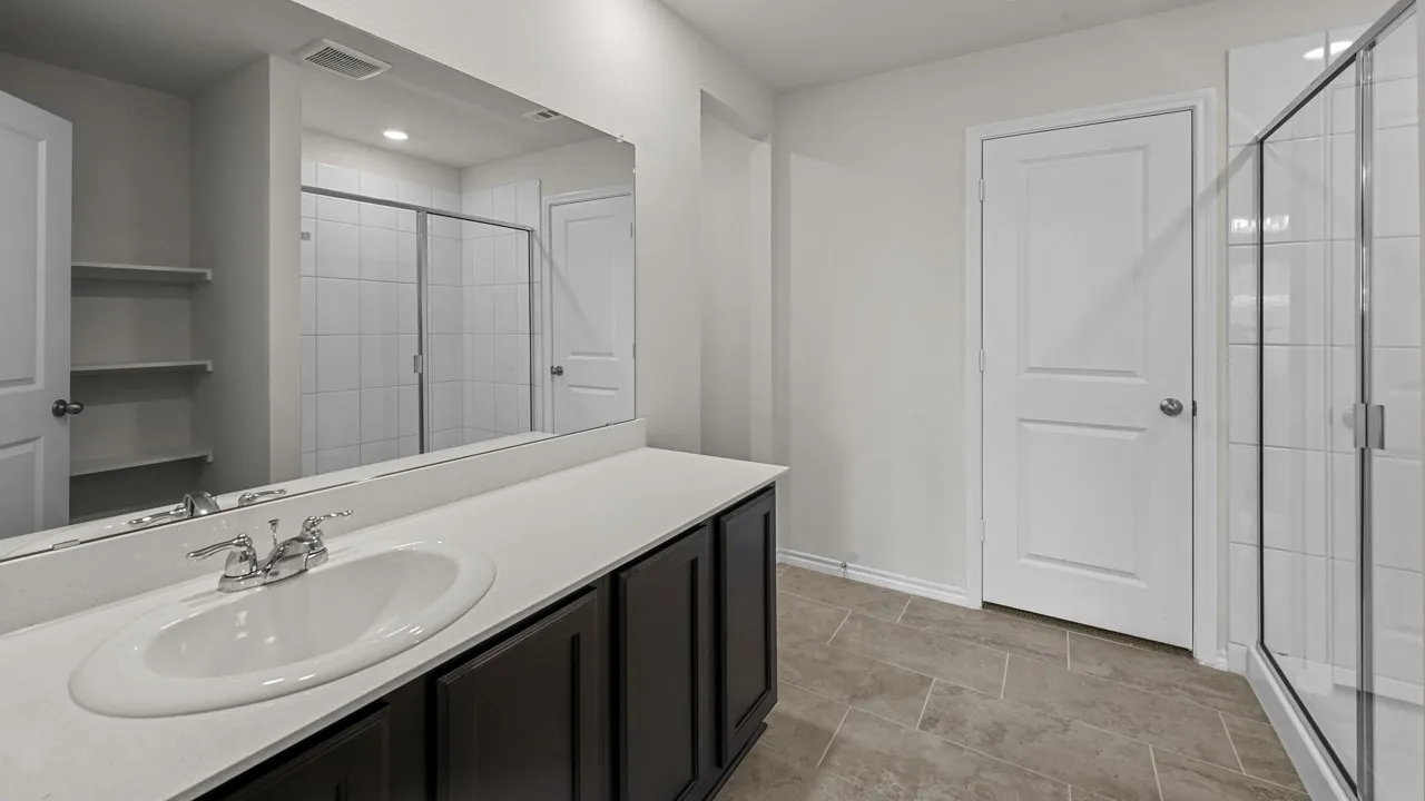 Bathroom featuring a white oval sink set in a long vanity with dark cabinetry, a large wall-mounted mirror, and a glass-enclosed shower with white tiled walls