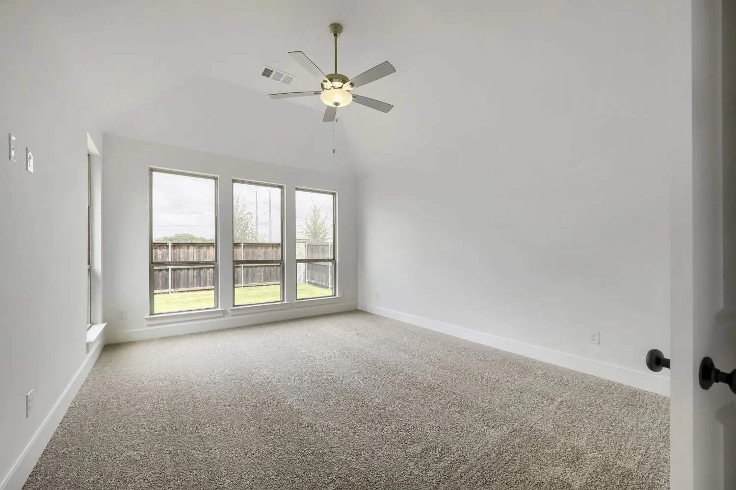 Carpeted interior space featuring a vaulted ceiling with a brass-finish ceiling fan and three large windows overlooking a fenced backyard