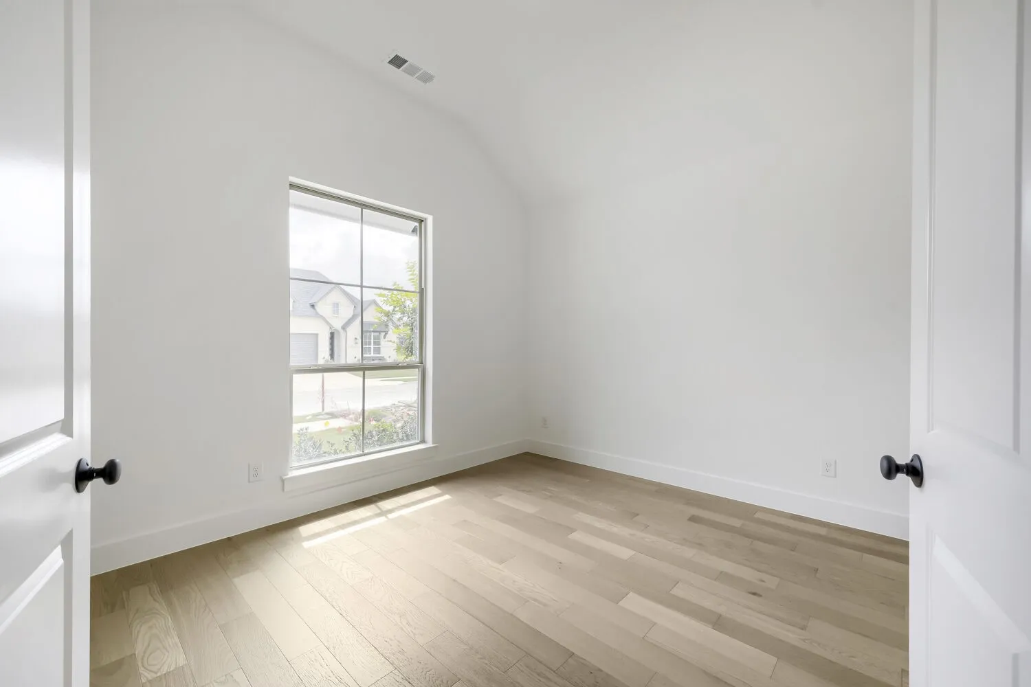Vaulted ceiling room featuring light wood-finish flooring, a multi-pane window with white trim, white walls, and a white door with a black knob