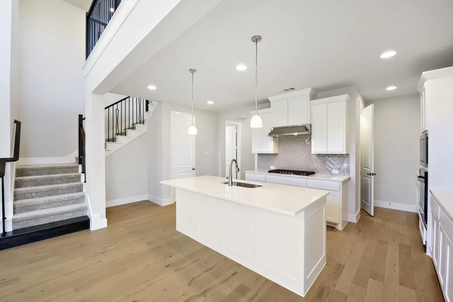 Open-concept living space with light wood-finish flooring, a two-story foyer, and a kitchen featuring a central island with sink, white cabinetry, and a herringbone tile backsplash