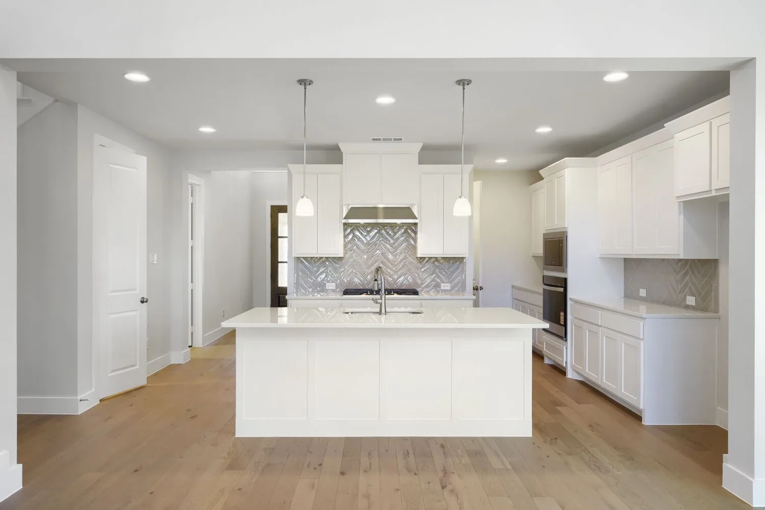 Contemporary kitchen featuring a large central island with pendant lighting, herringbone tile backsplash, integrated stainless steel appliances, white shaker cabinetry, and wood-finish flooring