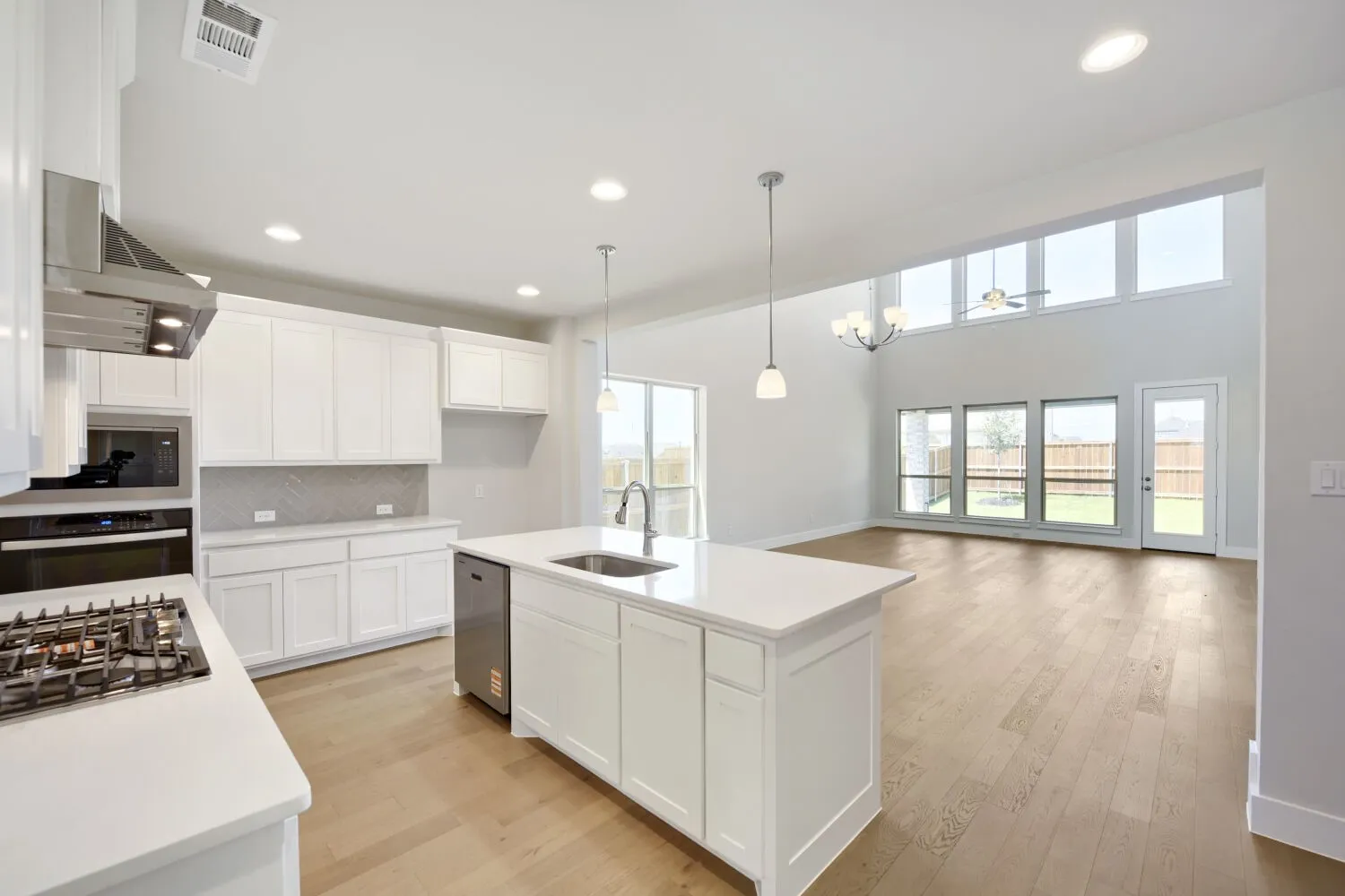 Bright open-concept kitchen featuring white shaker cabinetry, stainless steel appliances, a center island with sink, and light wood-finish flooring extending into the living area with a vaulted ceiling and transom windows