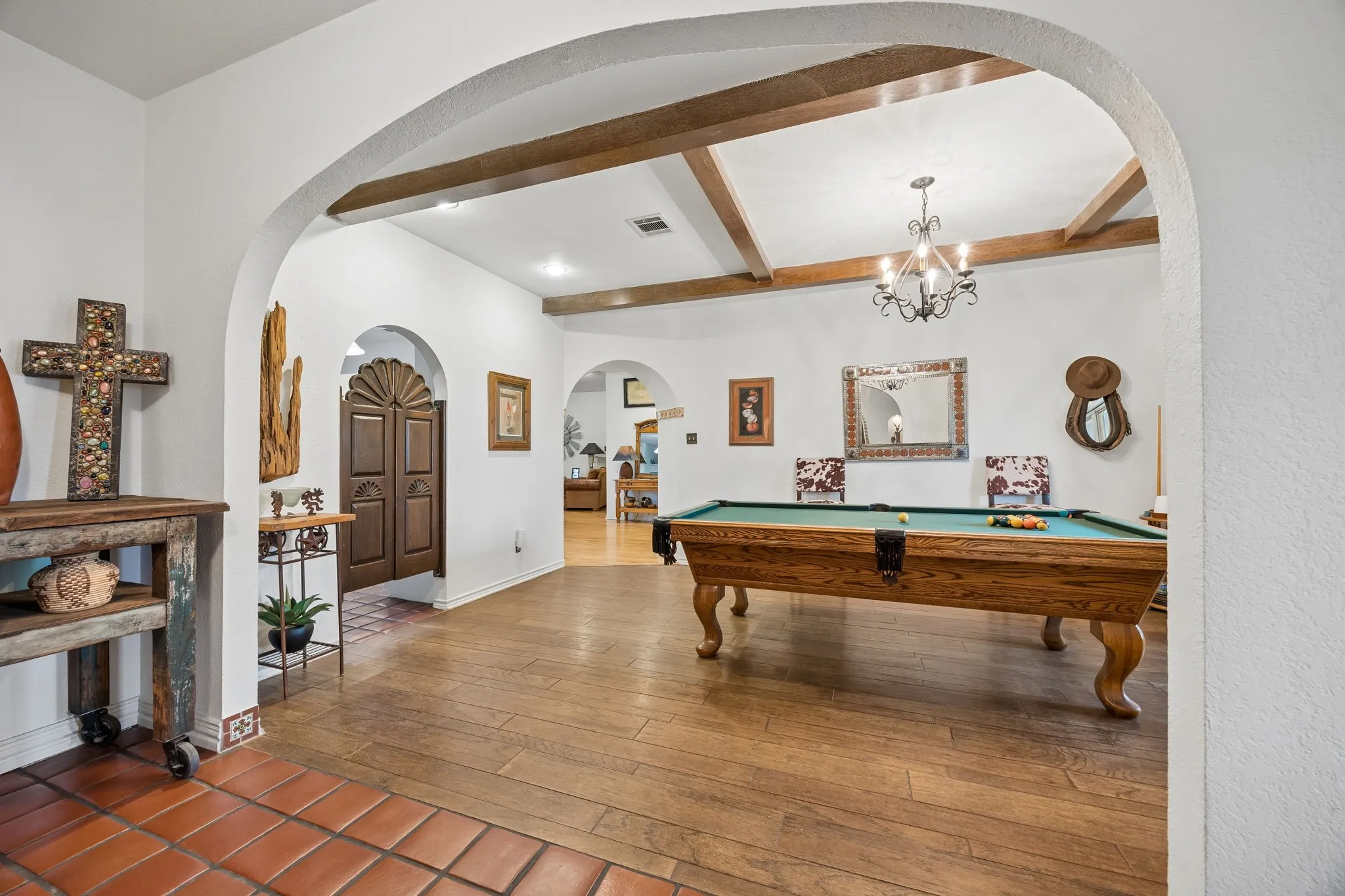 Arched entryway with terra cotta tile flooring opening to a living area featuring wood-finish flooring, exposed ceiling beams, and a wrought iron chandelier