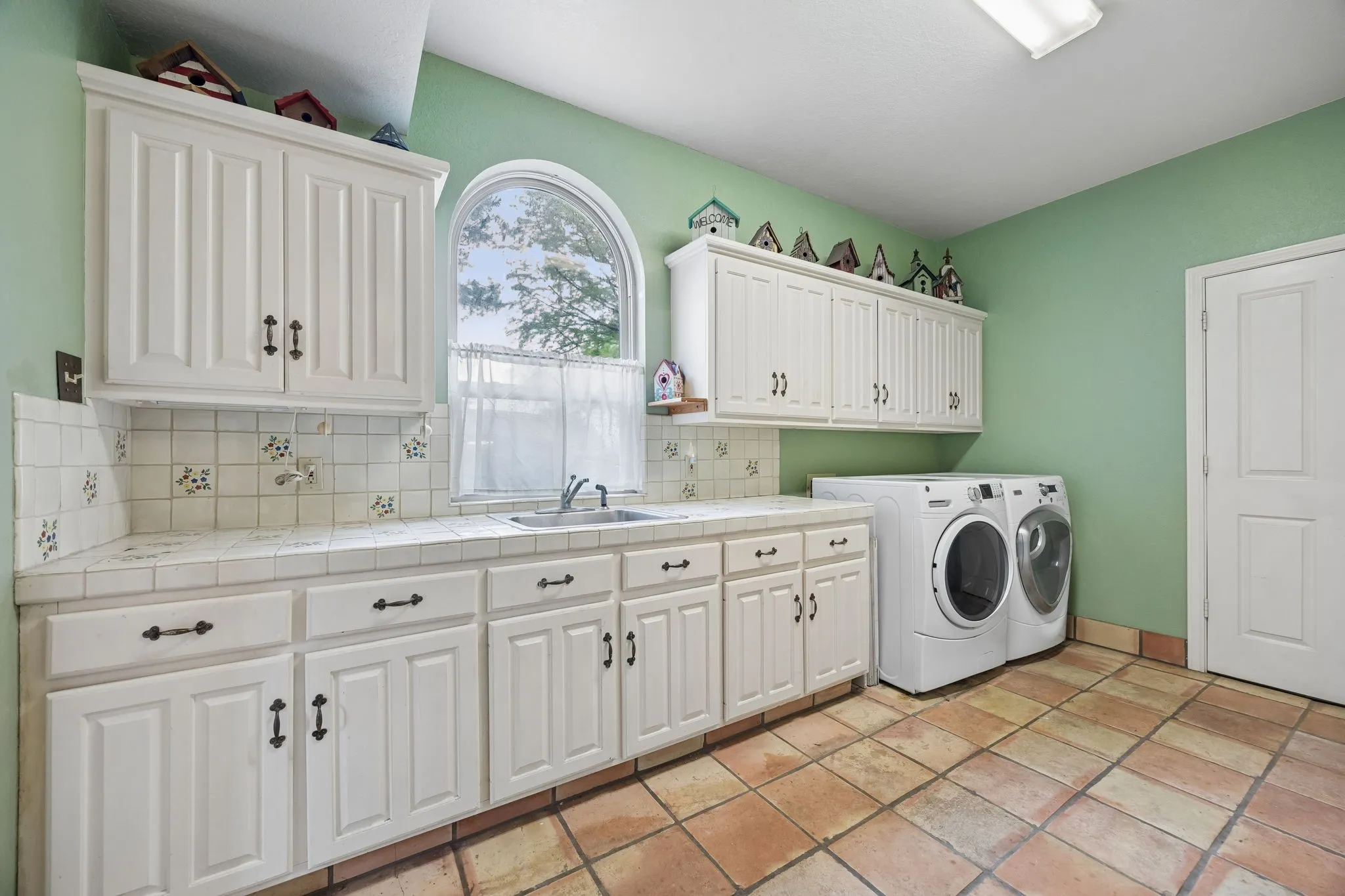 Laundry room featuring an arched window, ample cabinetry, ceramic tile countertops, a utility sink, and large format floor tiles