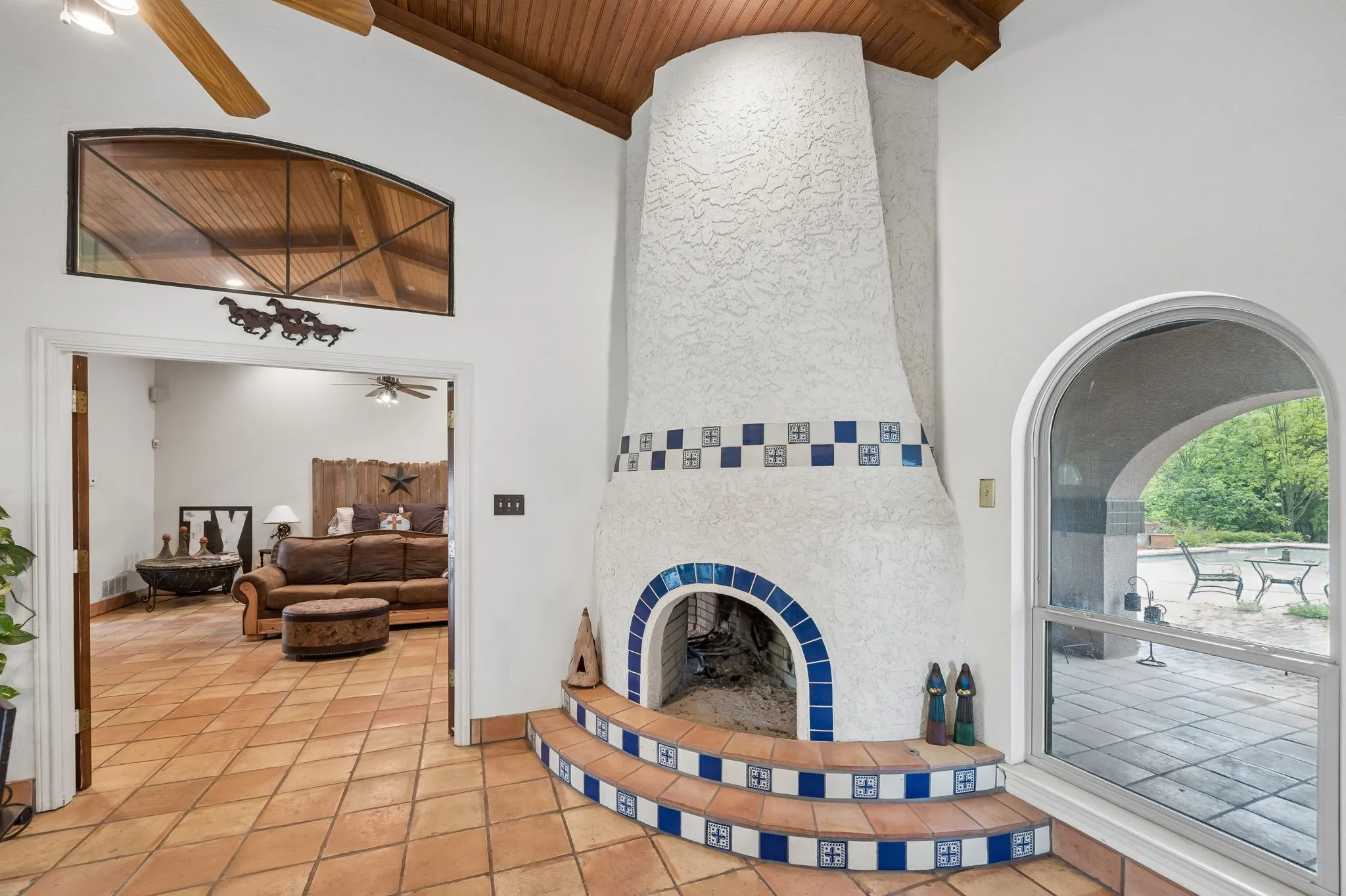 Exposed wood ceiling beams, a stucco fireplace with decorative blue and white tiling, and an arched window providing natural light