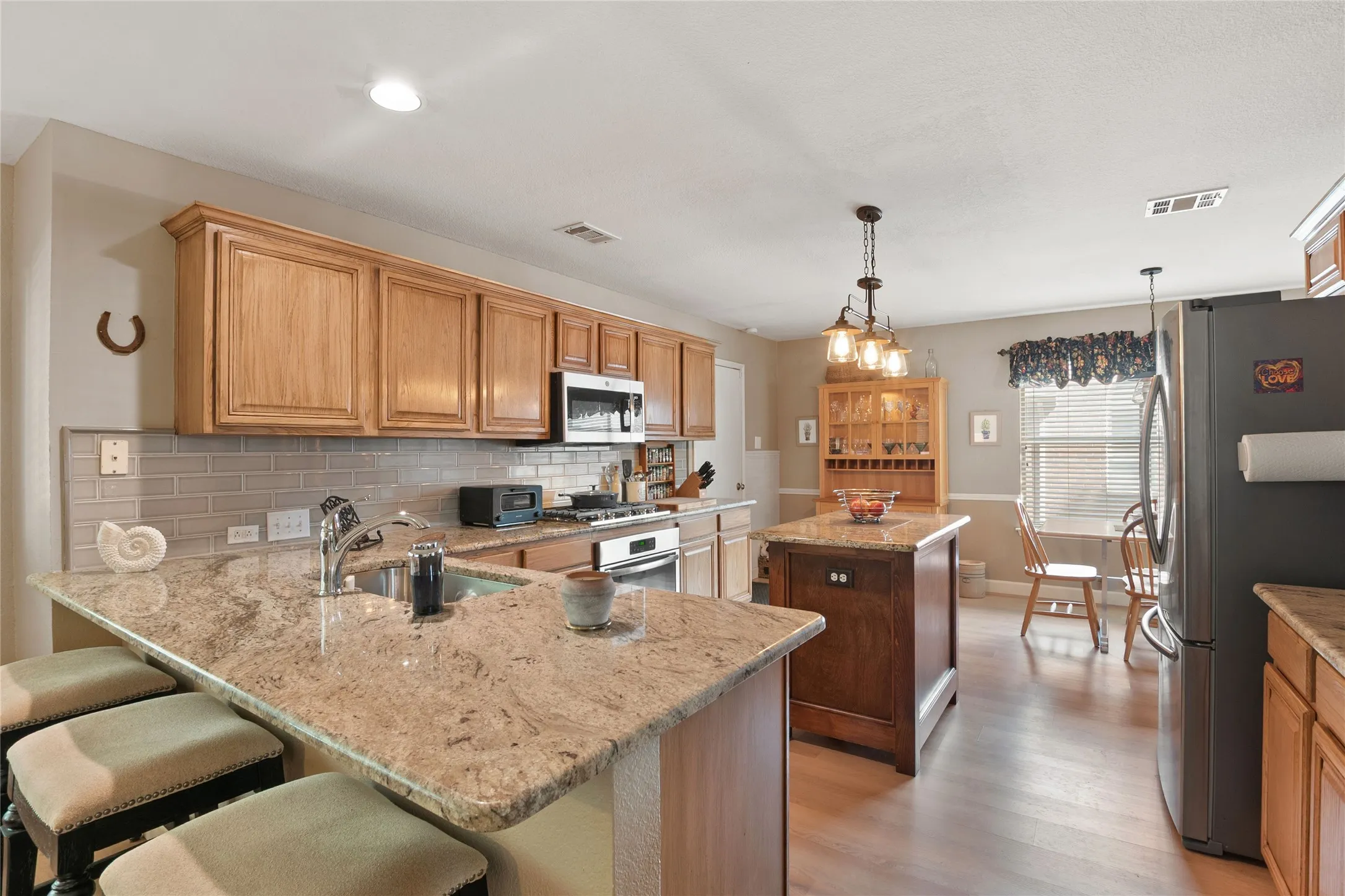 Kitchen featuring an island with a pendant light fixture, granite countertops, light wood-finish cabinetry, a subway tile backsplash, and stainless steel appliances