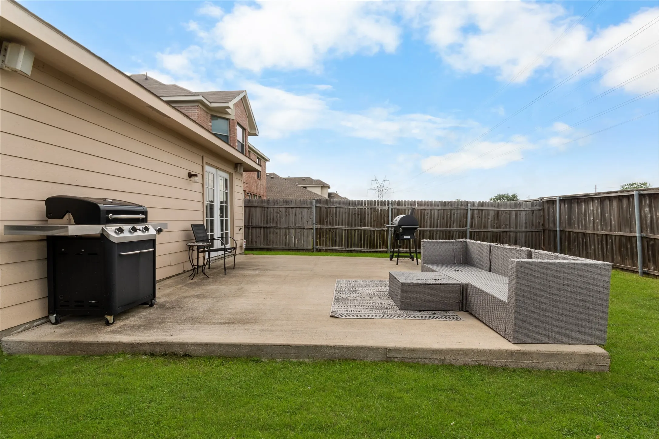 The property features a concrete patio area with direct access to the interior via double doors, surrounded by a green lawn and a wooden fence. Enhanced grass.