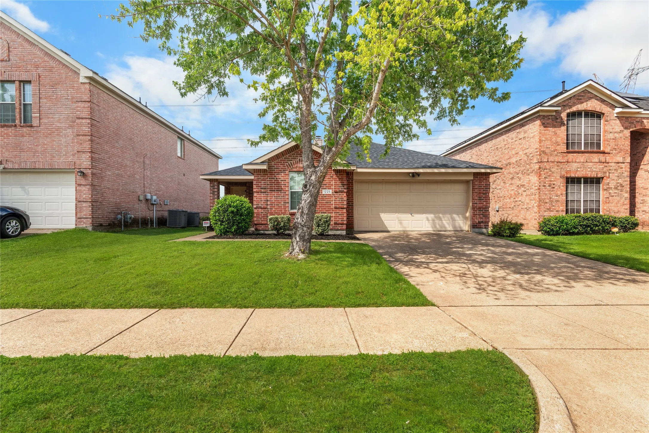 The property features a brick exterior, an attached garage, a paved driveway, and a well-maintained lawn with a mature tree. Enhanced grass.