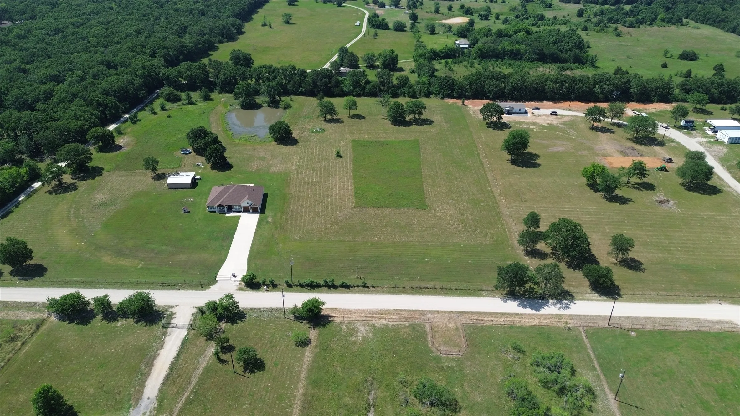 Expansive rural property featuring a single-story residence with a brown shingle roof, an extended concrete driveway, and a detached outbuilding