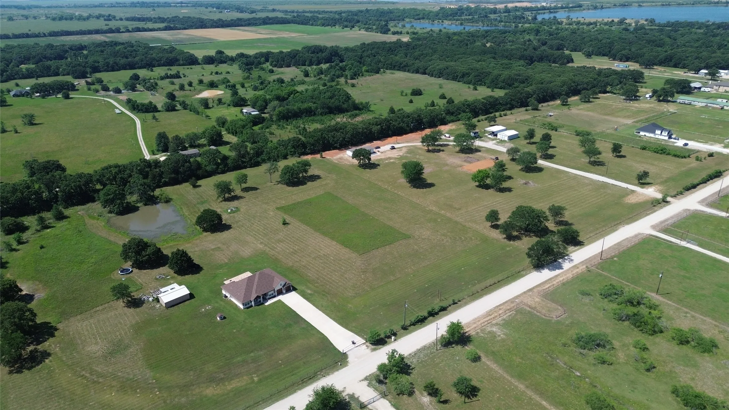 Expansive property featuring a single-story residence with a brown shingle roof, a long paved driveway, multiple outbuildings, and a pond