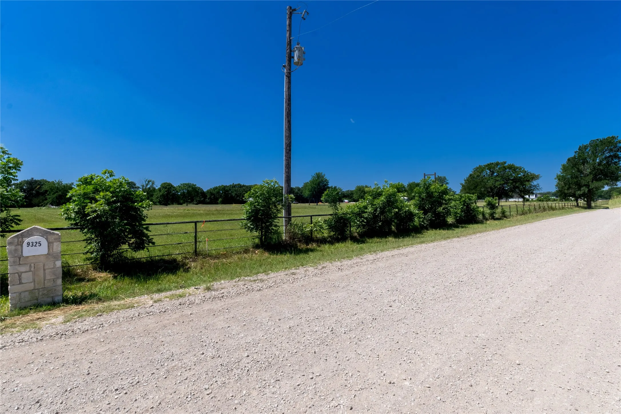 Gravel road frontage bordering a fenced pasture with mature trees