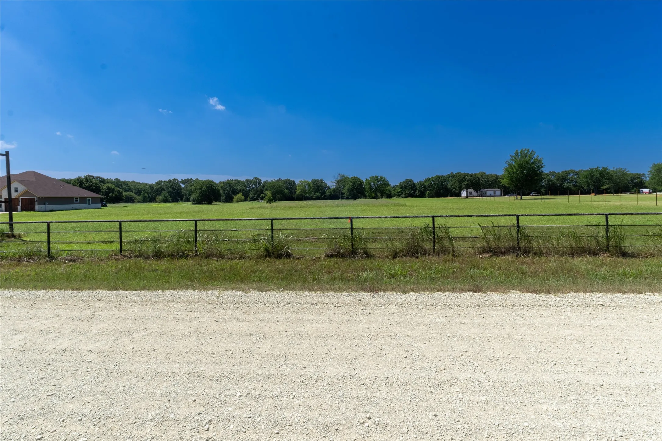 Expansive green acreage featuring a wire fence, mature trees, and a distant structure with a brown shingle roof