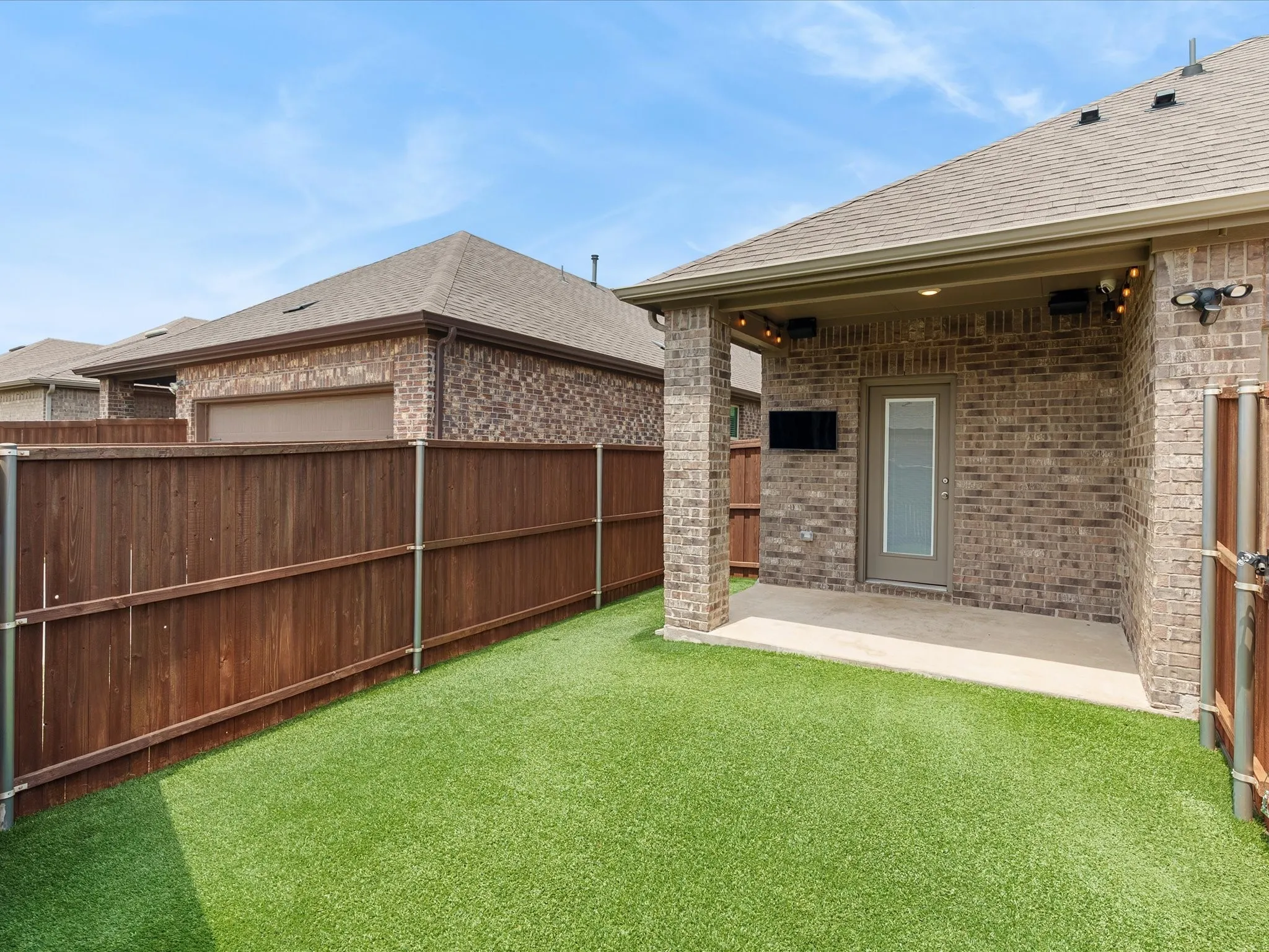 Covered patio and turf backyard complete with decorative lighting, built-in speakers, and mounted outdoor TV setup perfect for entertaining.