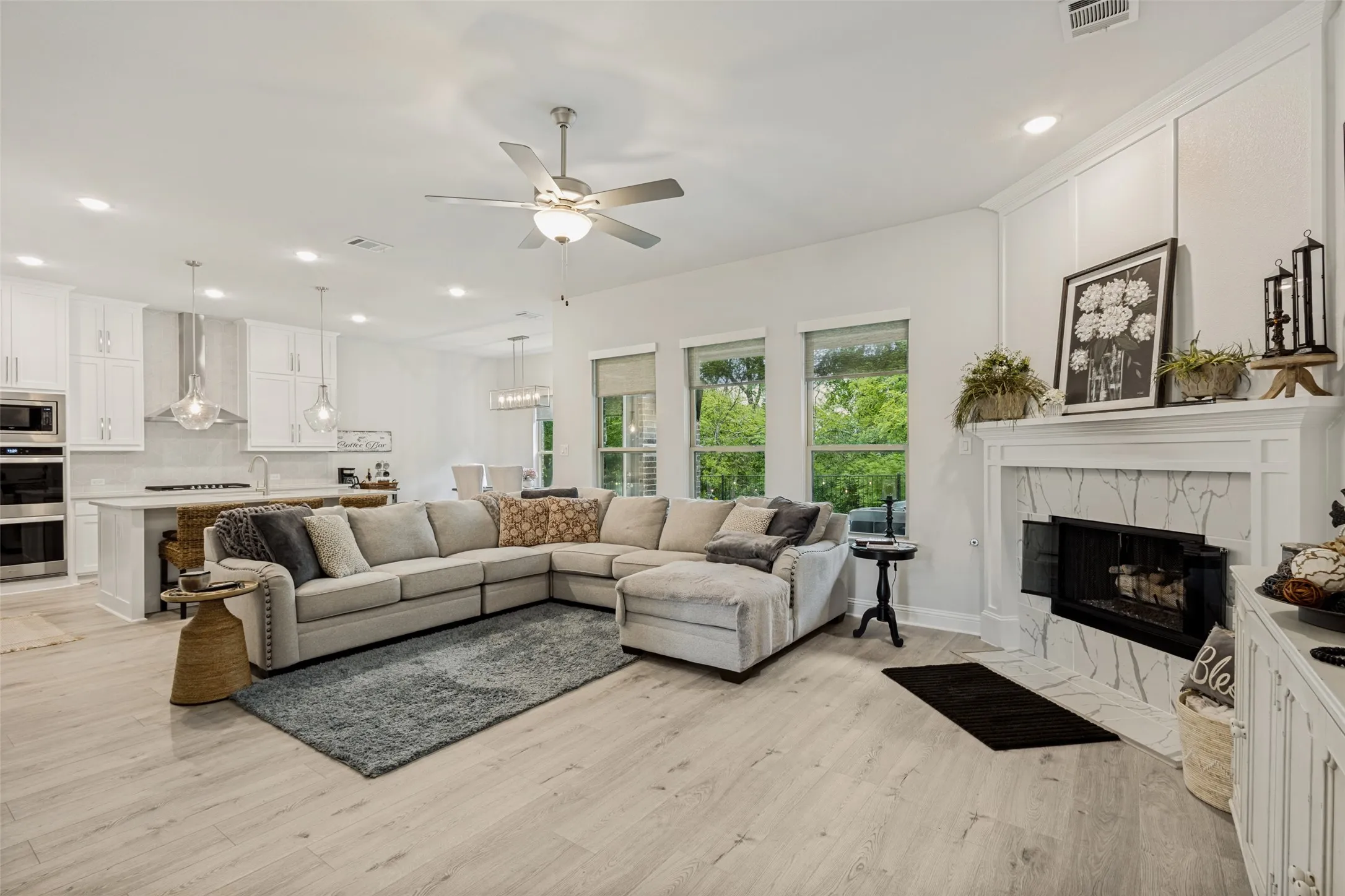 Spacious open concept living area featuring light-colored flooring, a ceiling fan, and a fireplace with a white mantel and marble-look surround