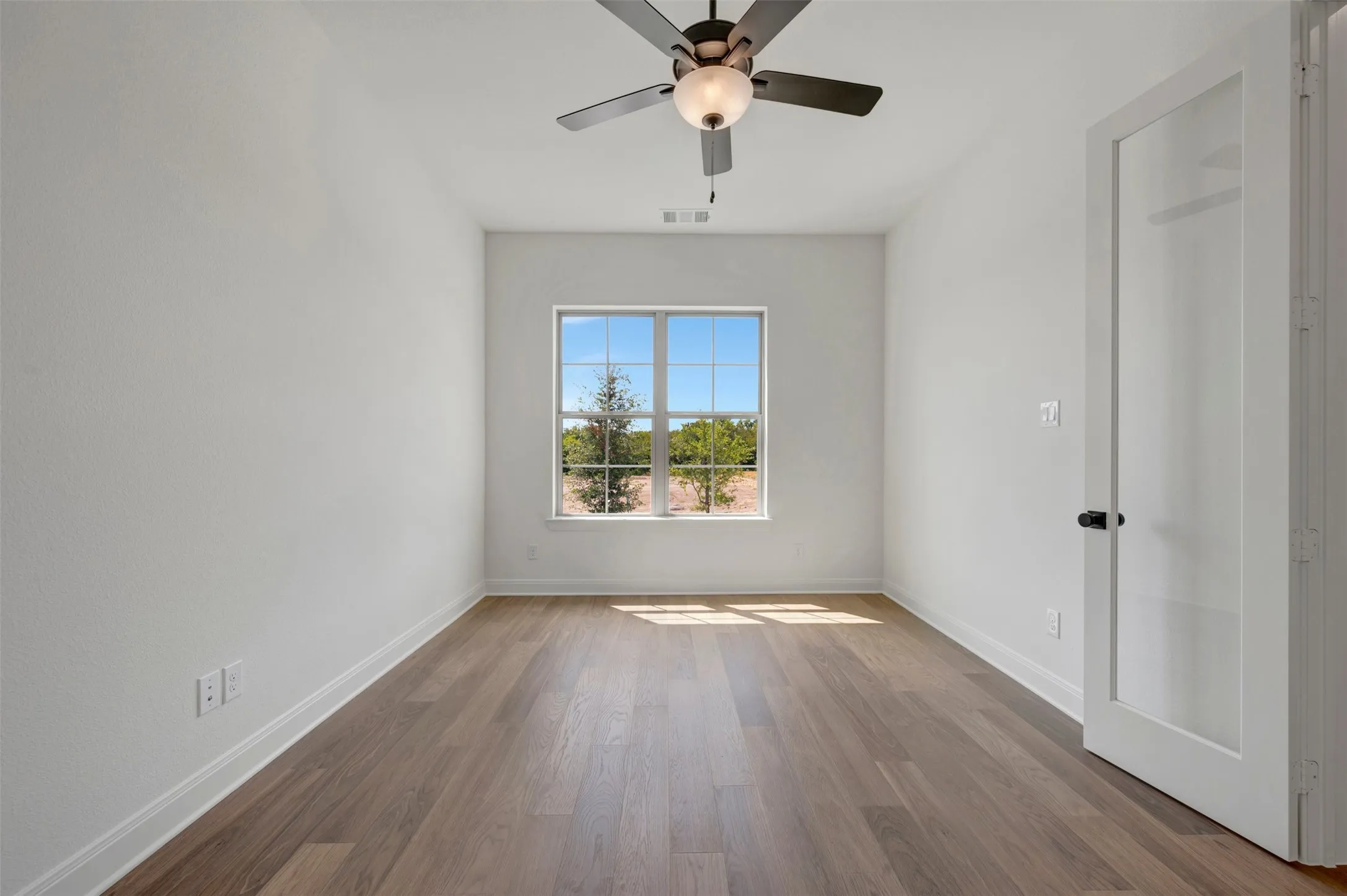 Wood-finish flooring, ceiling fan with integrated lighting, large window with outdoor scenery, and a white interior door with a matte black knob