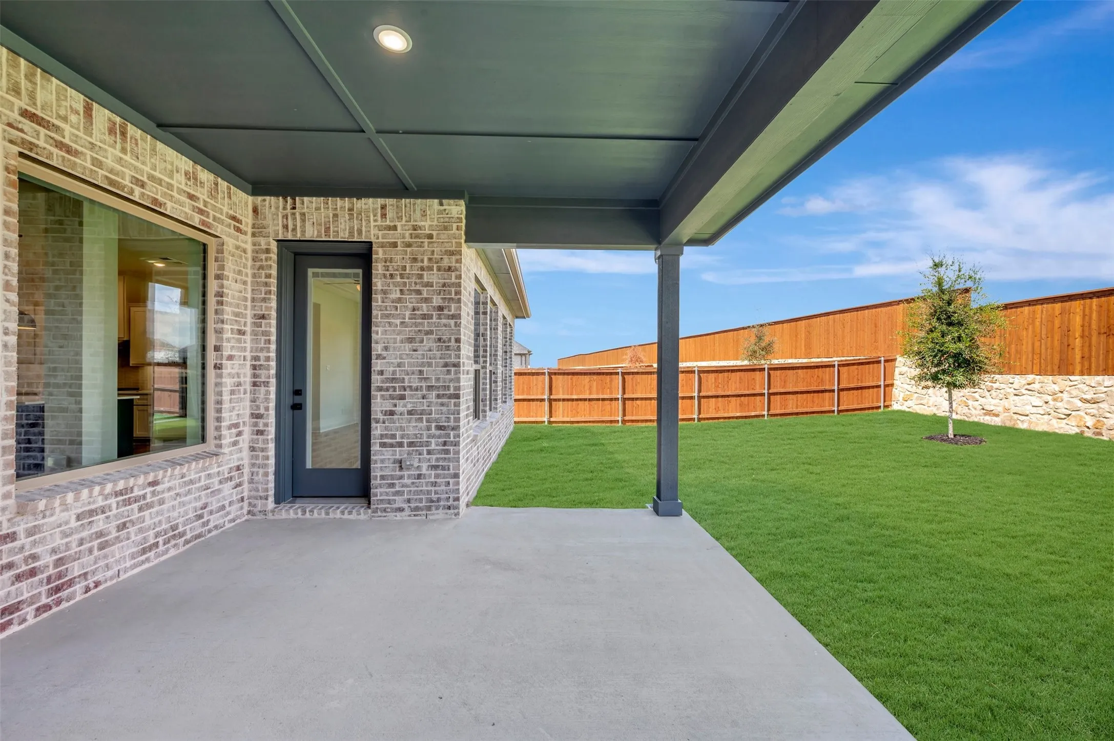 Covered patio with a concrete surface, a brick exterior, a manicured lawn, a wood fence, and a stone retaining wall