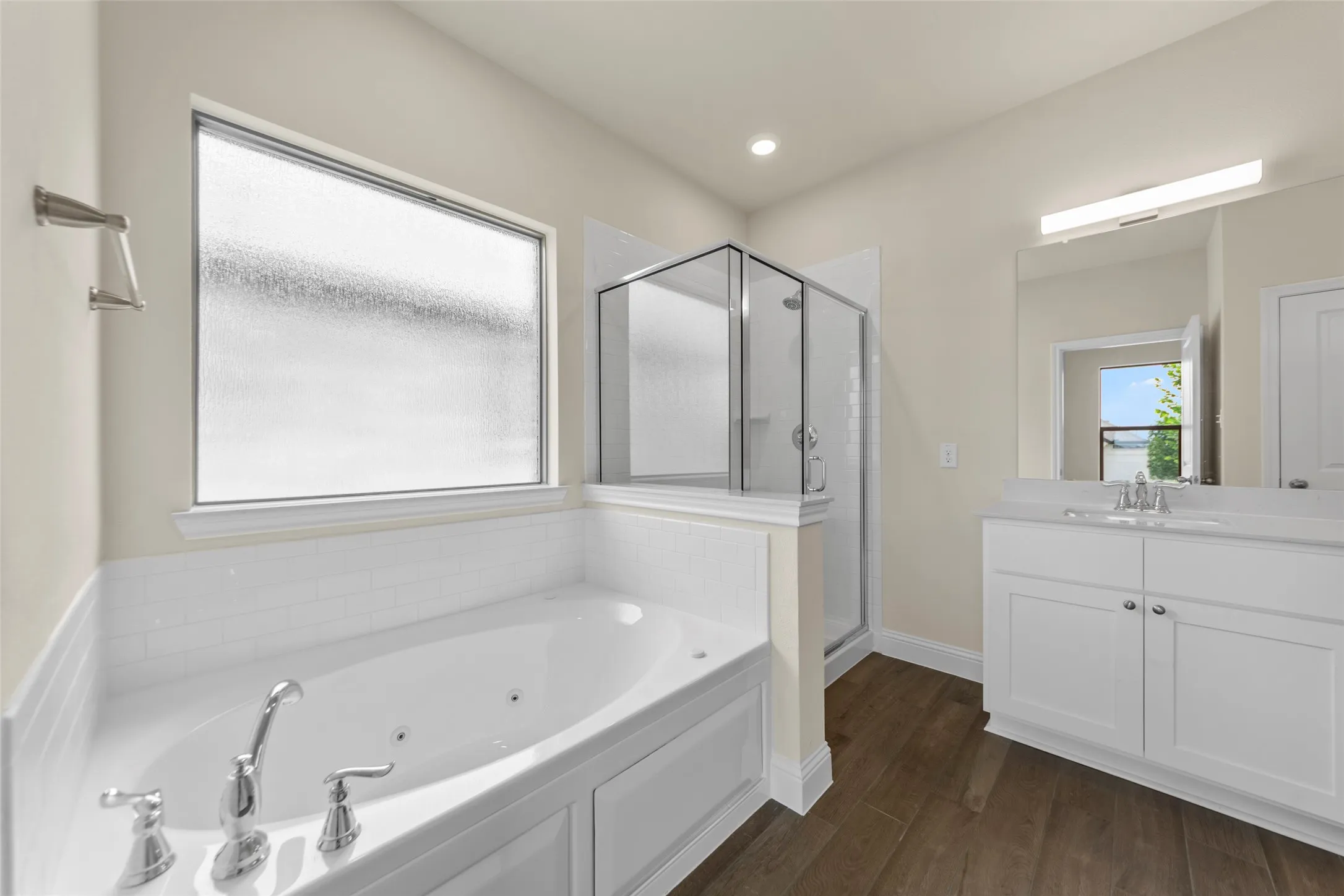 Bathroom featuring a soaking tub with polished chrome fixtures, a separate glass-enclosed shower, wood-finish flooring, a white vanity with an integrated sink, and an expansive mirror