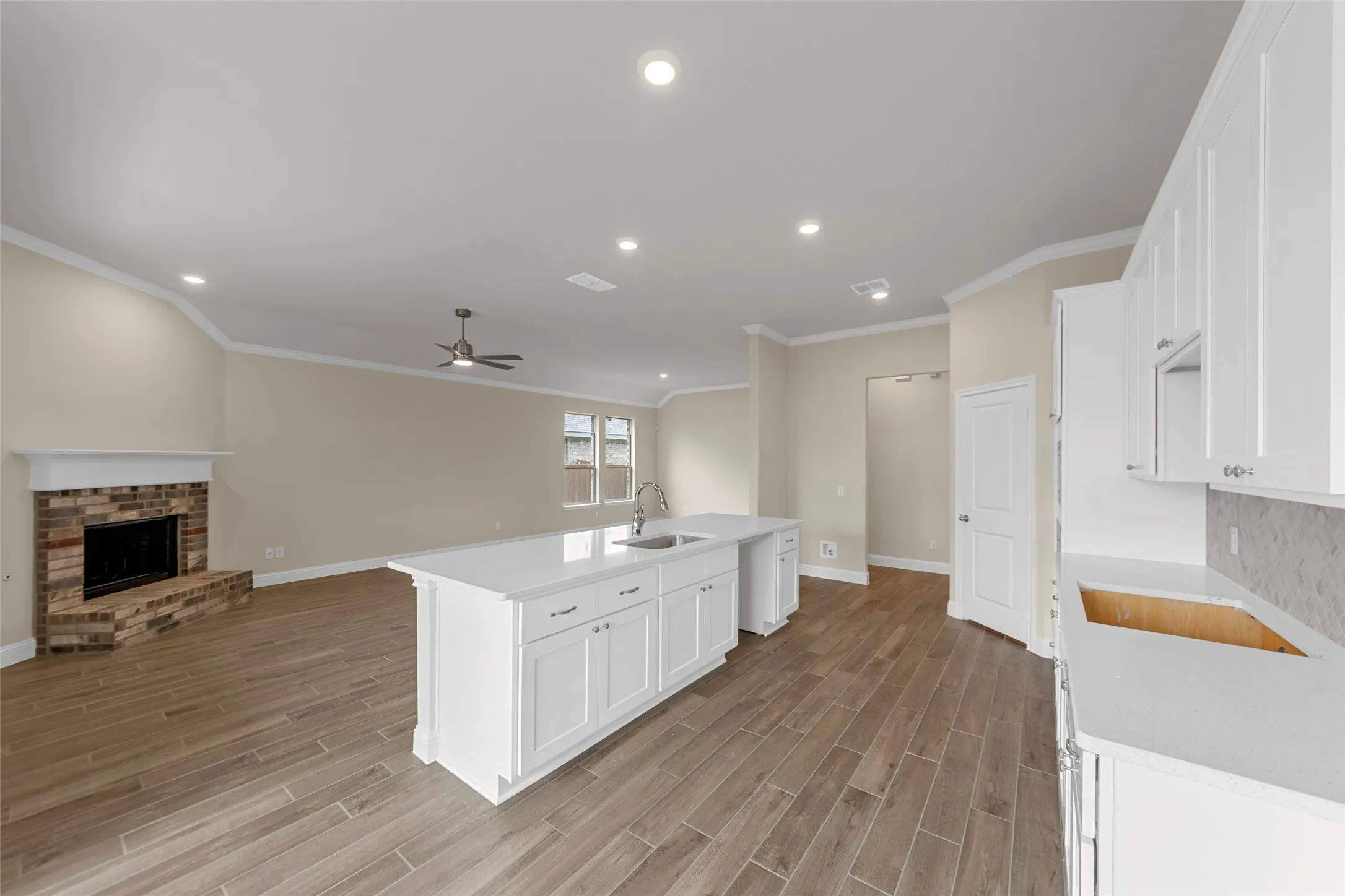 Open-concept living area featuring wood-finish flooring, a kitchen island with integrated sink, white cabinetry with stone countertops, a brick fireplace, and recessed lighting