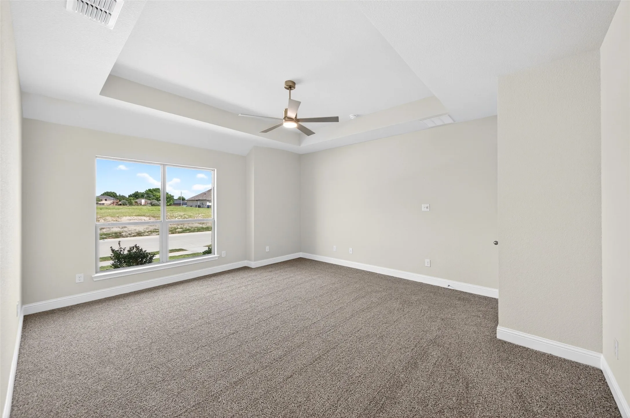 Spacious interior room featuring a tray ceiling with a ceiling fan, light gray wall paint, gray carpet flooring, and a large window providing ample natural illumination