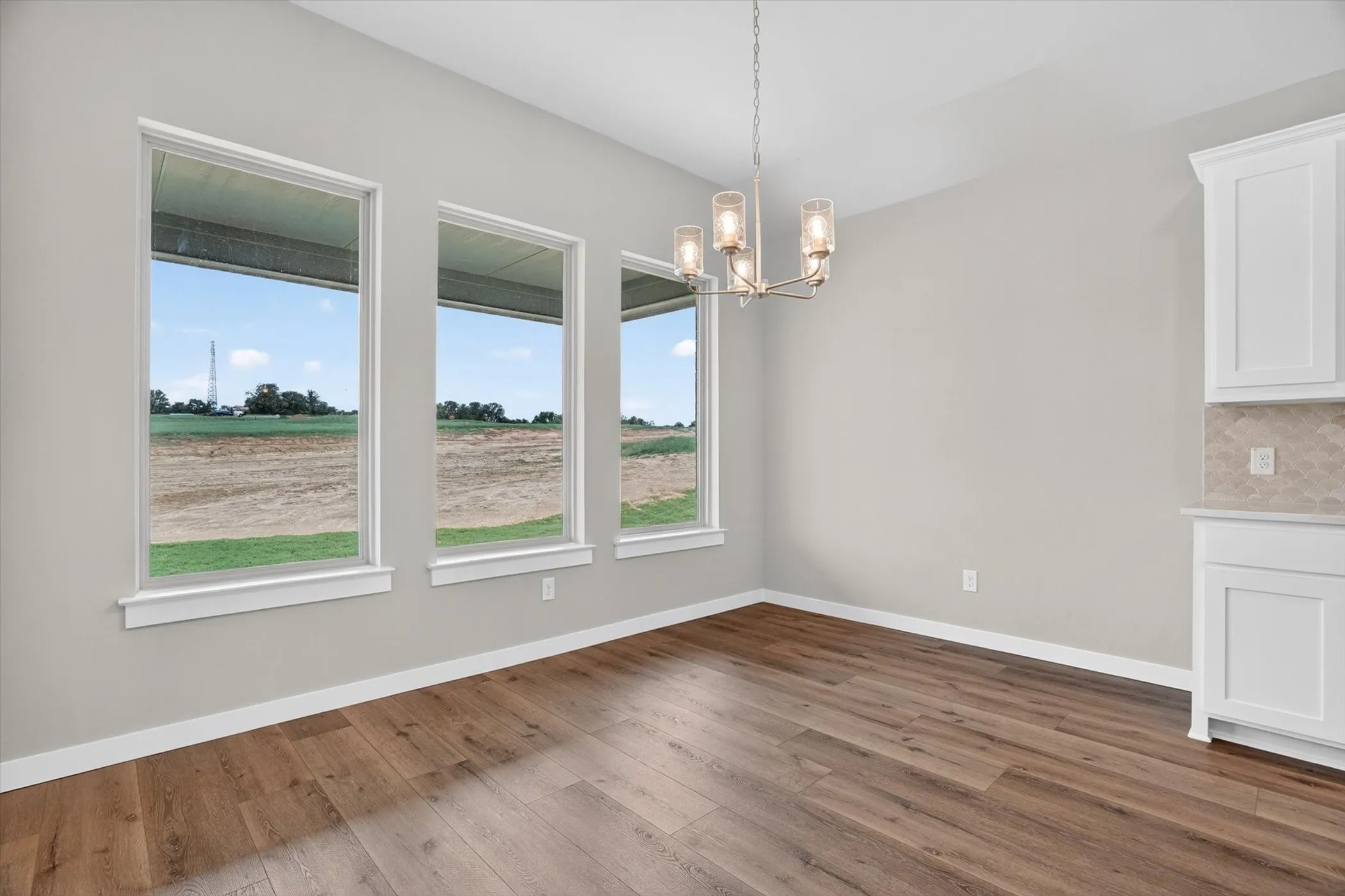 Interior space featuring three rectangular windows, wood-finish flooring, a contemporary chandelier, white baseboards, and built-in white cabinetry with a patterned backsplash