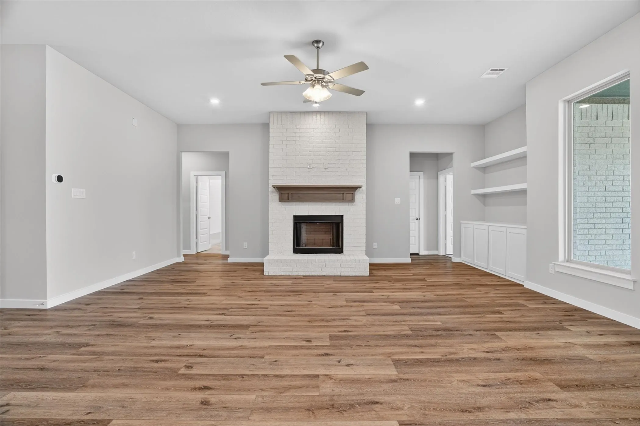 Spacious living area featuring wood-finish flooring, a white brick fireplace with a wood mantle, built-in shelving, white cabinetry, and a ceiling fan