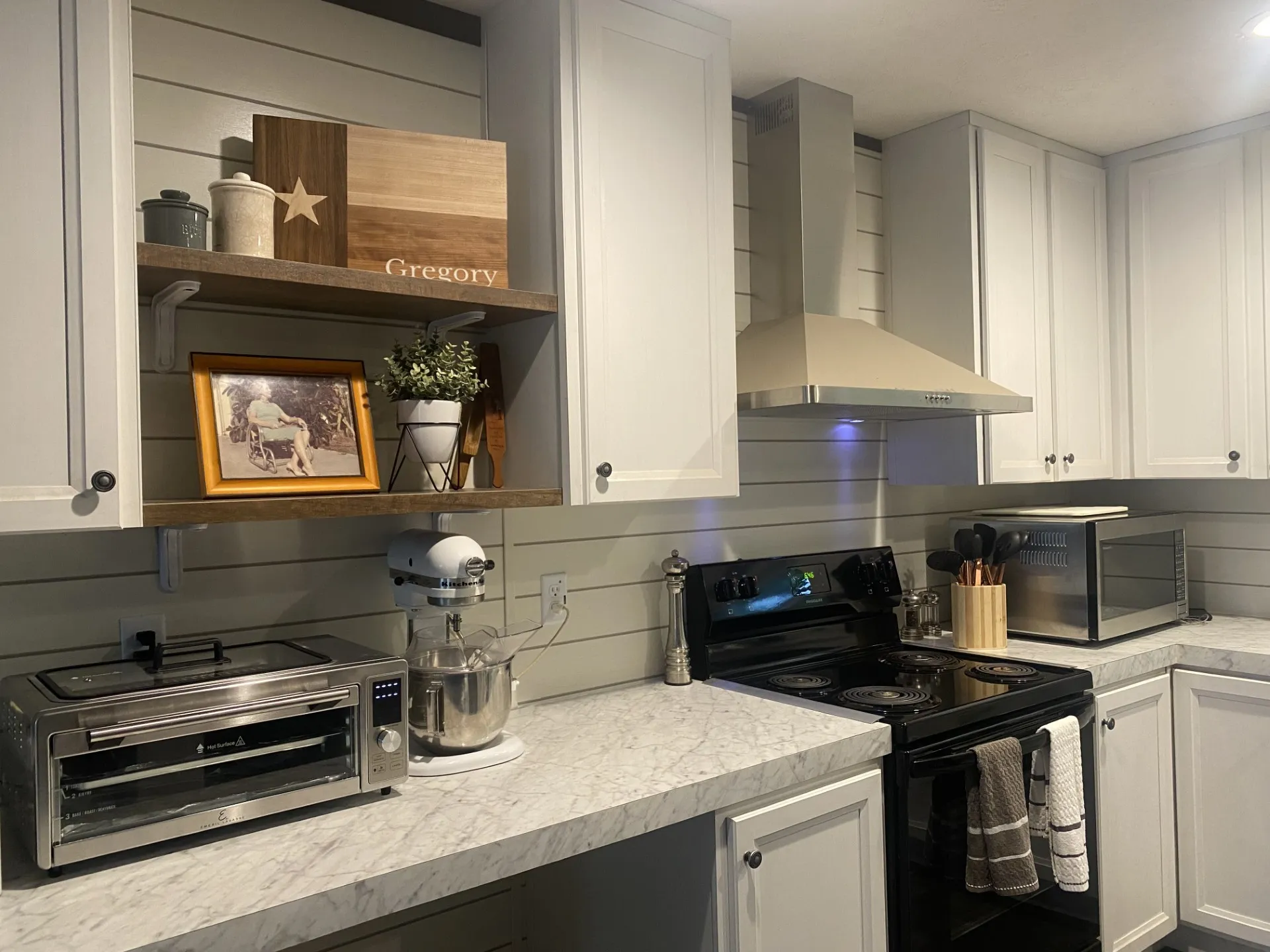 Kitchen featuring white shaker-style cabinetry, a stainless steel range hood, a black electric range, open shelving with dark wood finish, and light-toned countertops