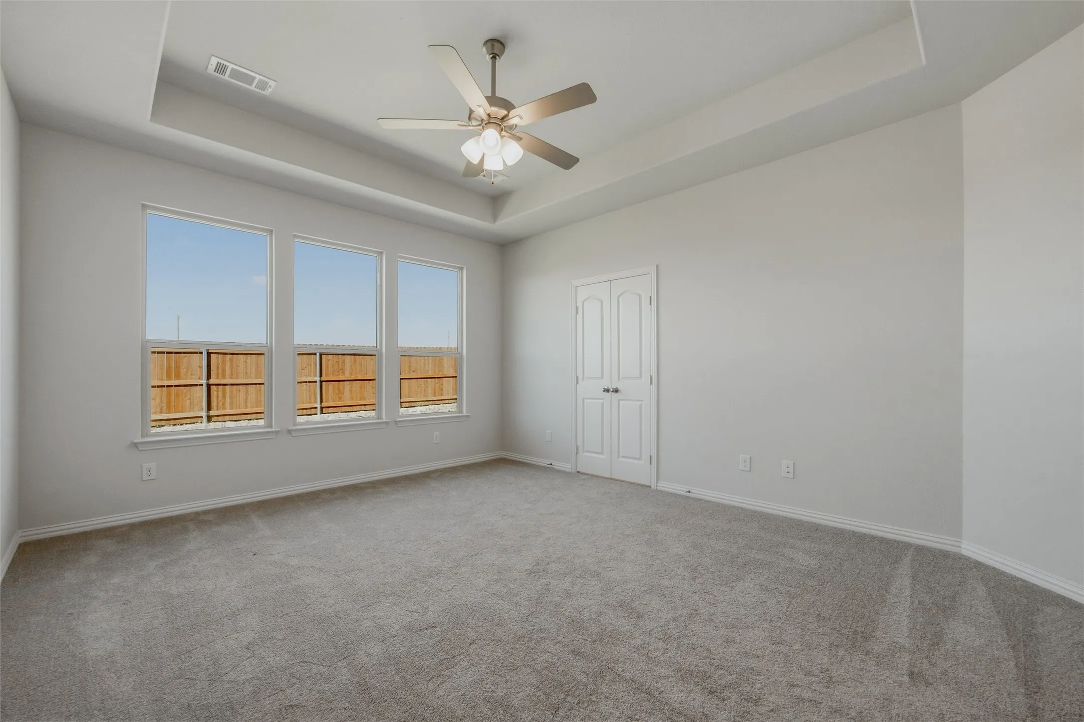Tray ceiling, ceiling fan with light kit, three large windows, gray carpeting, and double panel door