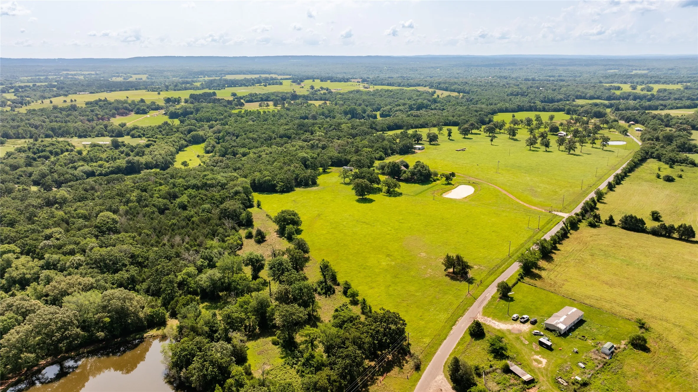 Expansive acreage featuring a mix of open fields, mature tree lines, a pond, and a winding road