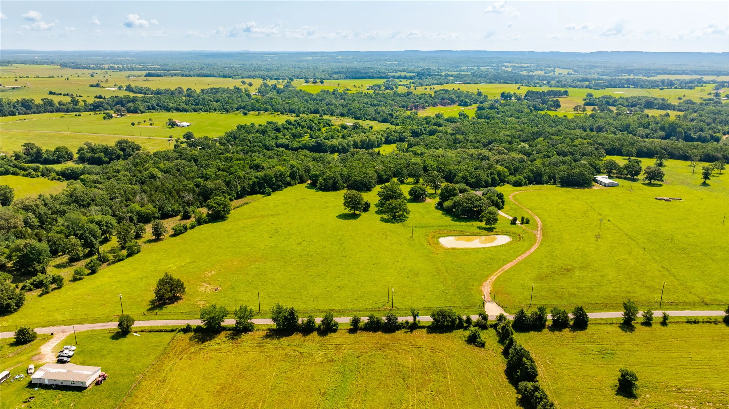 Expansive green acreage featuring wooded areas, open fields, a pond, and a winding dirt road