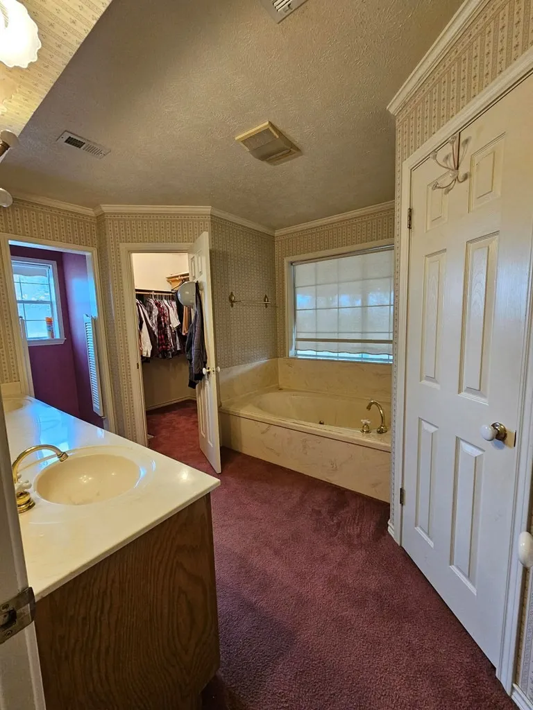 Full bath featuring vanity, wallpapered walls, ornamental molding, a textured ceiling, and dark colored carpet
