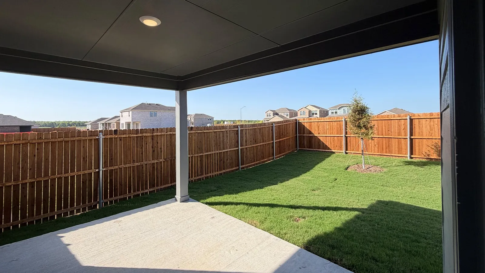 Covered patio with recessed lighting, concrete flooring, and a private wood fence enclosing a lawn with a young tree