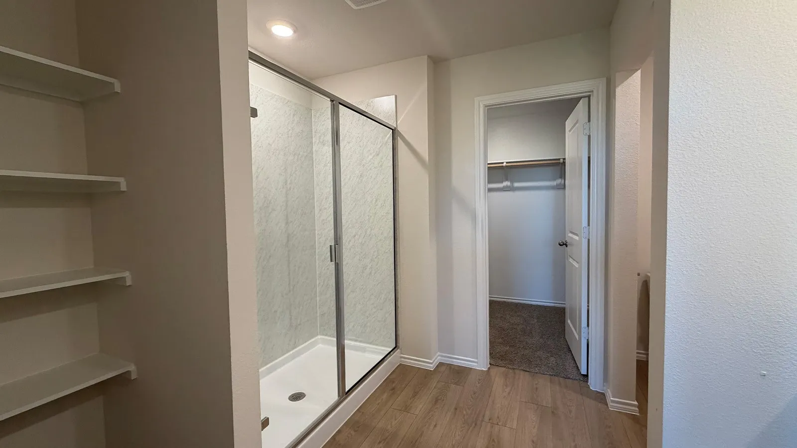 Walk-in shower with glass enclosure and white base, featuring light-toned wood-finish flooring, recessed lighting, and open shelving