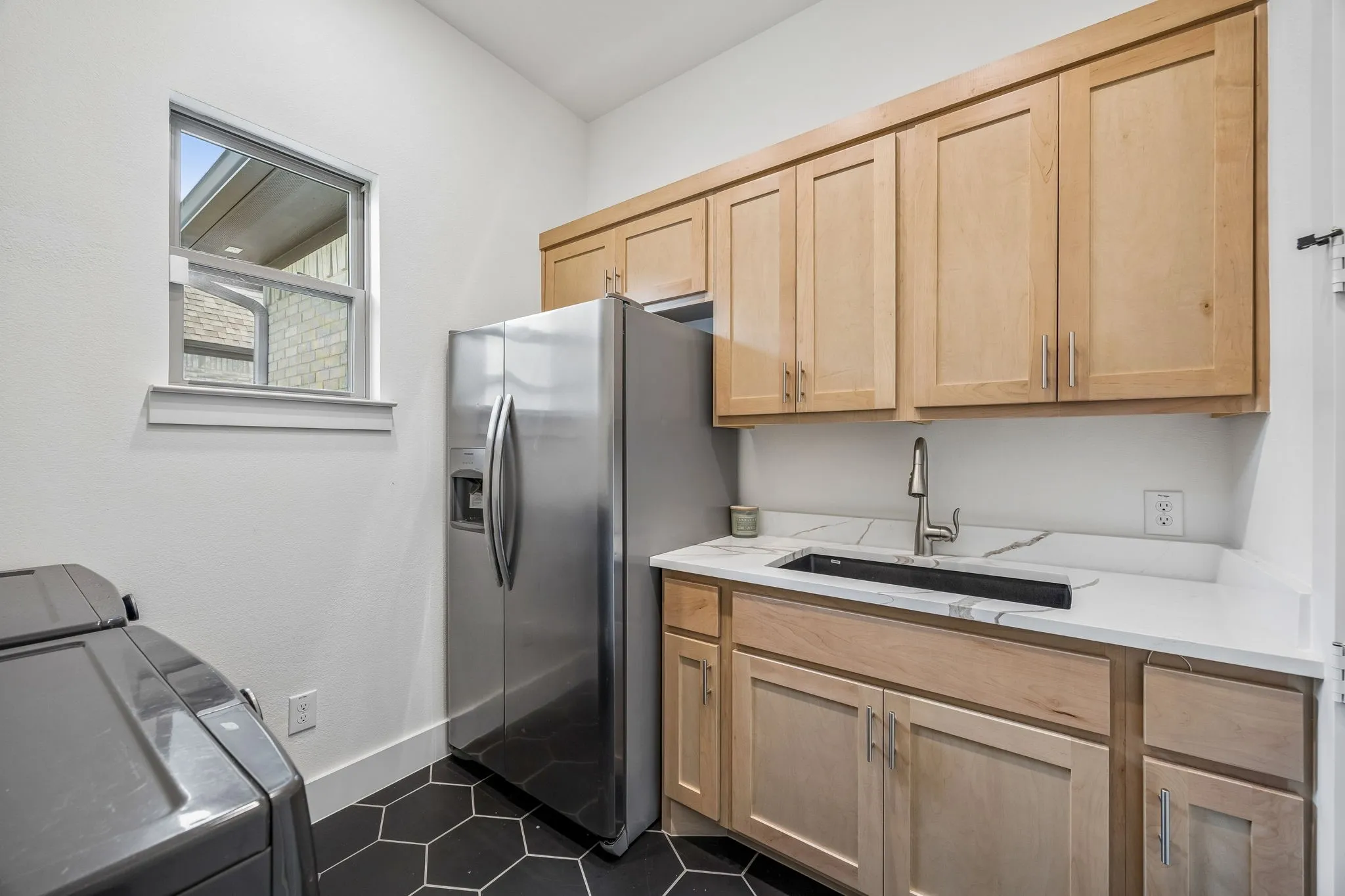 Kitchen with light wood finish cabinetry, stainless steel fridge with ice dispenser, dark tile patterned flooring, and washing machine and dryer