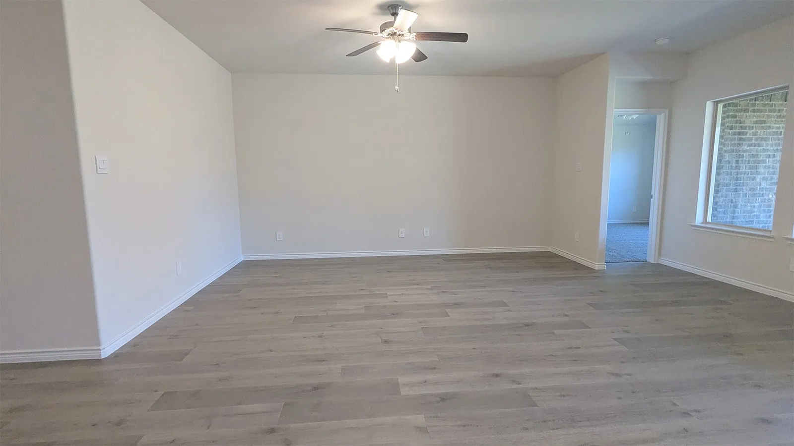 Spacious room featuring light wood-finish flooring, neutral wall tones, a ceiling fan with integrated lighting, and a large window providing natural illumination