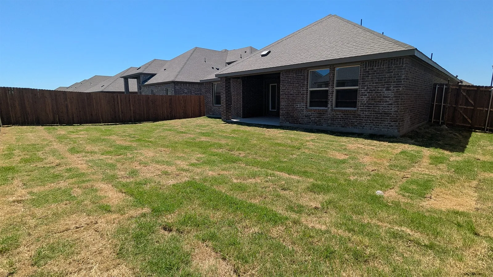 Expansive backyard with a full privacy fence, a concrete patio, and a brick exterior home featuring multiple windows and a shingled roof