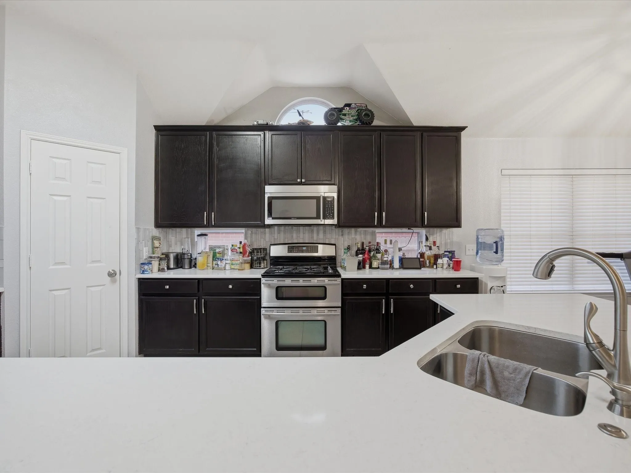 Kitchen featuring stainless steel appliances, dark wood finish cabinetry, light stone counters, vaulted ceiling, and tasteful backsplash