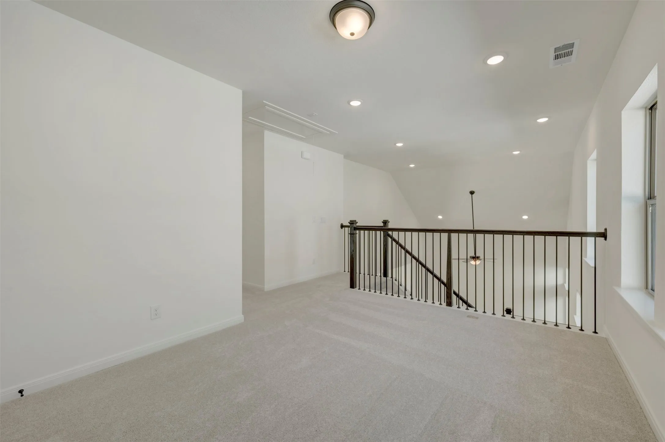 Spacious loft area featuring light gray carpeting, white walls, multiple recessed lights, a ceiling-mounted light fixture, and an iron baluster railing overlooking the floor below
