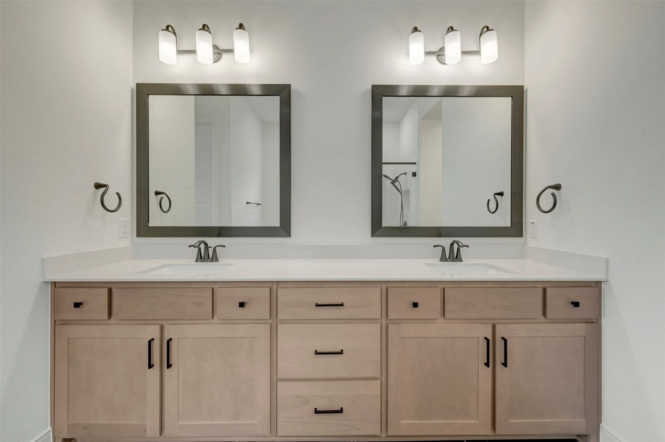Dual vanity featuring undermount sinks, light wood-finish cabinetry, matte black hardware, white countertops, and framed mirrors with brushed nickel light fixtures