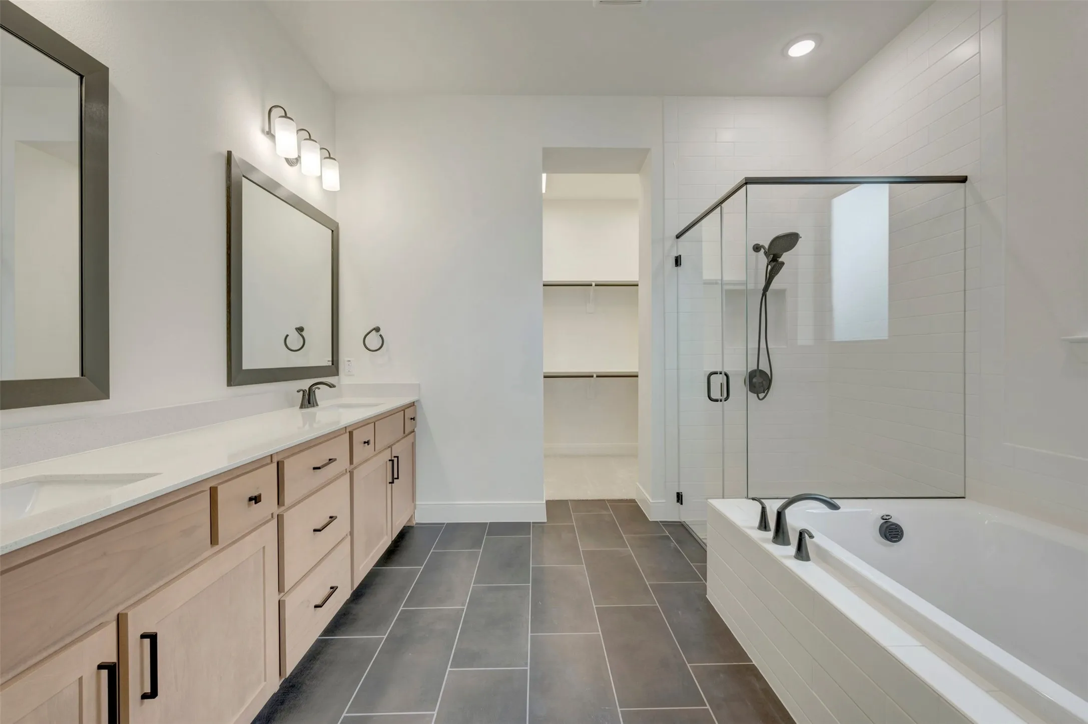 Spacious bathroom featuring a dual vanity with light wood-finish cabinetry and white countertops, a walk-in shower with a glass enclosure, a built-in soaking tub, and large format tile flooring