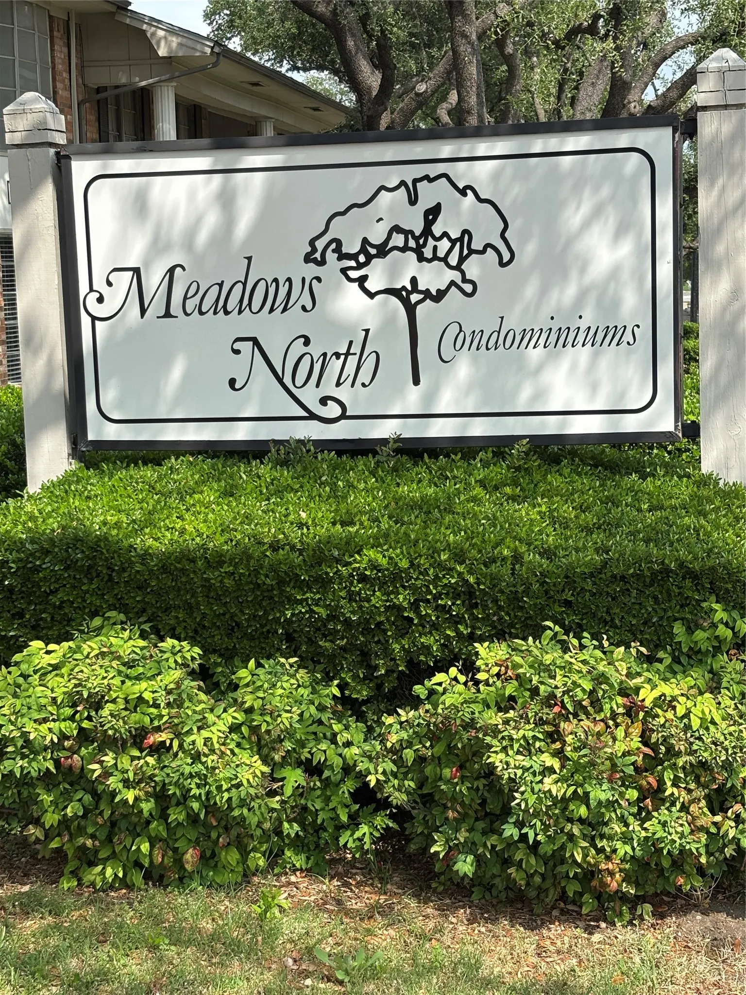 The property is part of Meadows North Condominiums, featuring a prominent welcome sign surrounded by lush green shrubs and mature trees