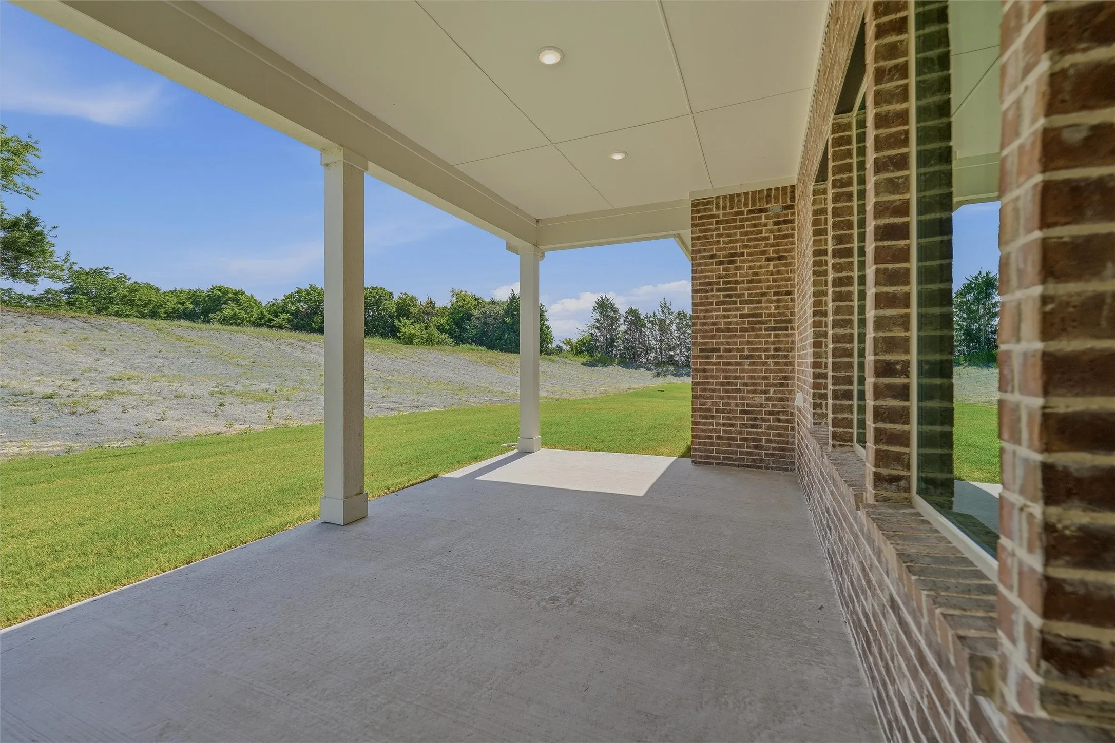 Covered concrete patio featuring recessed lighting, white support columns, and a brick exterior wall with multiple windows