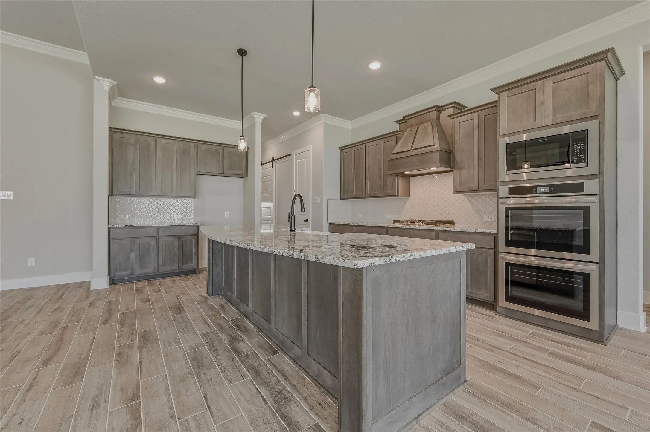 Gourmet kitchen featuring a large curved island with a stone countertop, wood-finish flooring, built-in stainless steel appliances, custom wood cabinetry, and a geometric tile backsplash